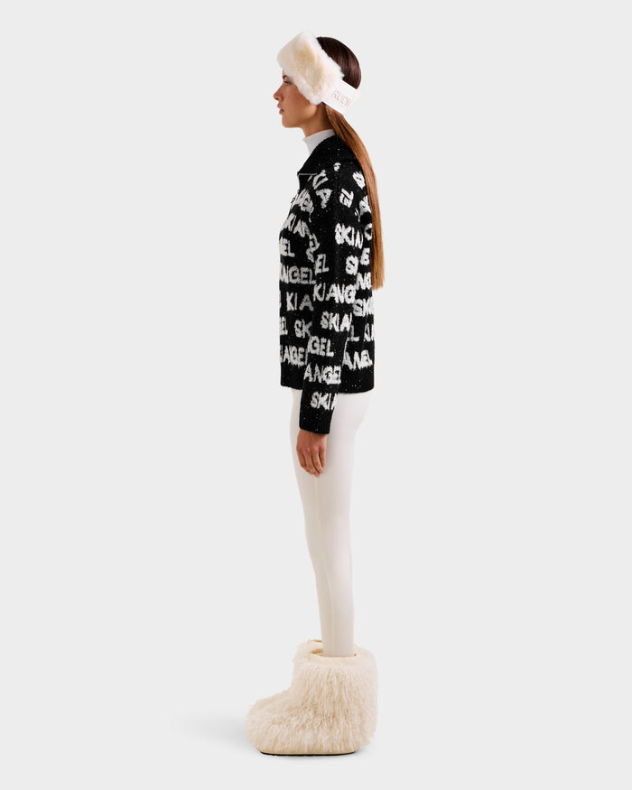 A woman in profile wears the Rudsak LYDIA - 8525687 Black women’s ski sweater, paired with white leggings, a fluffy white headband, and boots, against a plain light background.