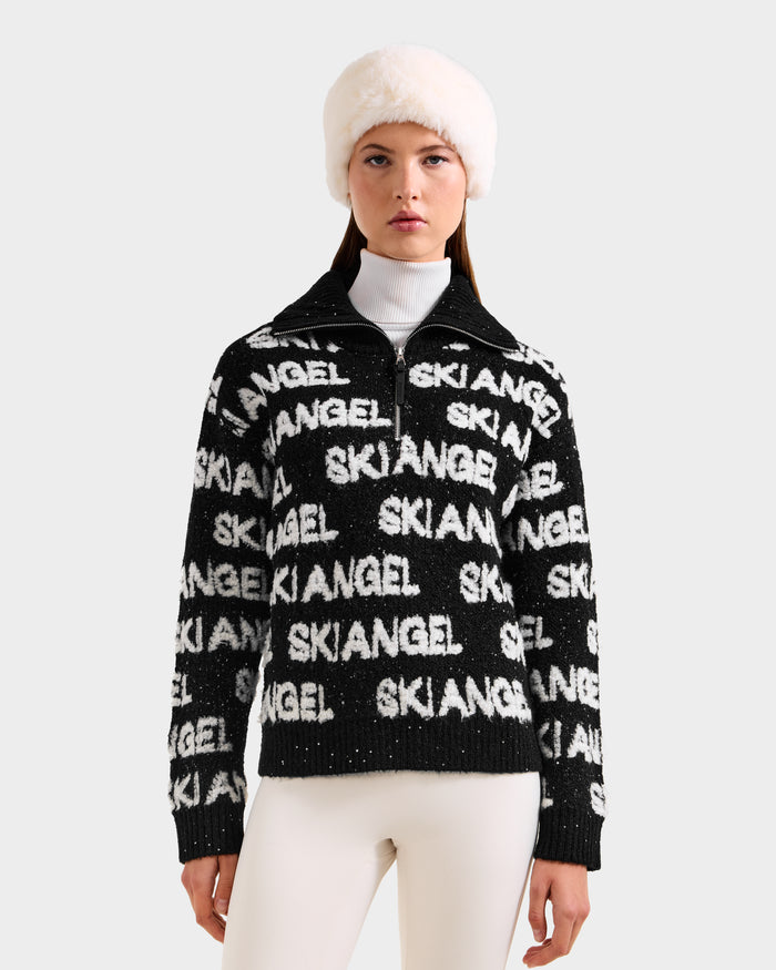 A woman confidently poses against a light background in white pants, a white furry hat, and the Rudsak LYDIA - 8525687 Black après-ski pullover over a white turtleneck.