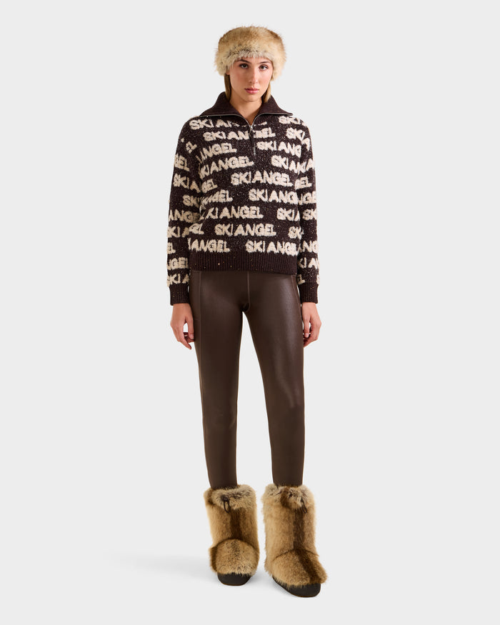 A person faces forward wearing Rudsak's LYDIA - 8525687 Espresso sweater, brown leggings, a furry hat, and tan fur boots—showcasing cozy après-ski style against a plain light background.