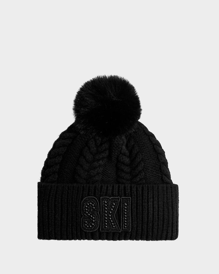 The Rudsak MAE - 8425550 Black is a black cable knit tuque with a large faux fur pom pom and "SKI" embroidered in black with embellishments on the folded brim.