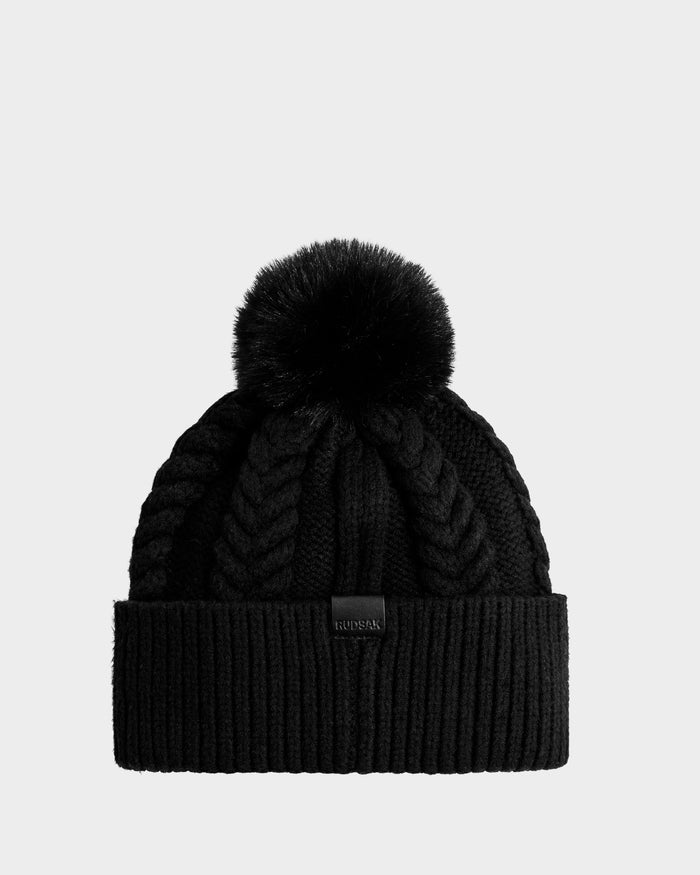 The Rudsak MAE - 8425550 Black is a women's cable knit tuque with a ribbed fold-over cuff, chunky braided patterns, a large faux fur pom pom, and a small rectangular front tag.