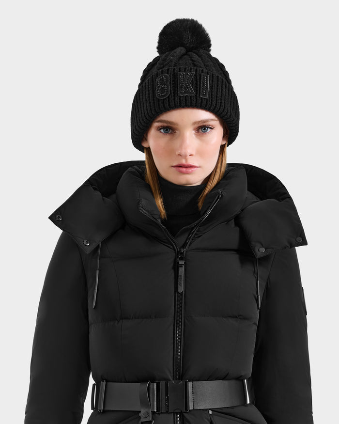 A woman stands against a light gray background wearing the Rudsak MAE - 8425550 Black puffer jacket with a belt and a women's cable knit tuque featuring a crystal-embellished SKI logo.