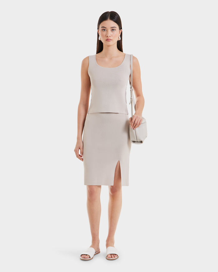 A woman stands facing forward, wearing the Rudsak MARIE - L525007 Cream minimalist knit top and matching knee-length skirt with a front slit. She carries a light beige shoulder bag and wears white slide sandals against a plain white background.