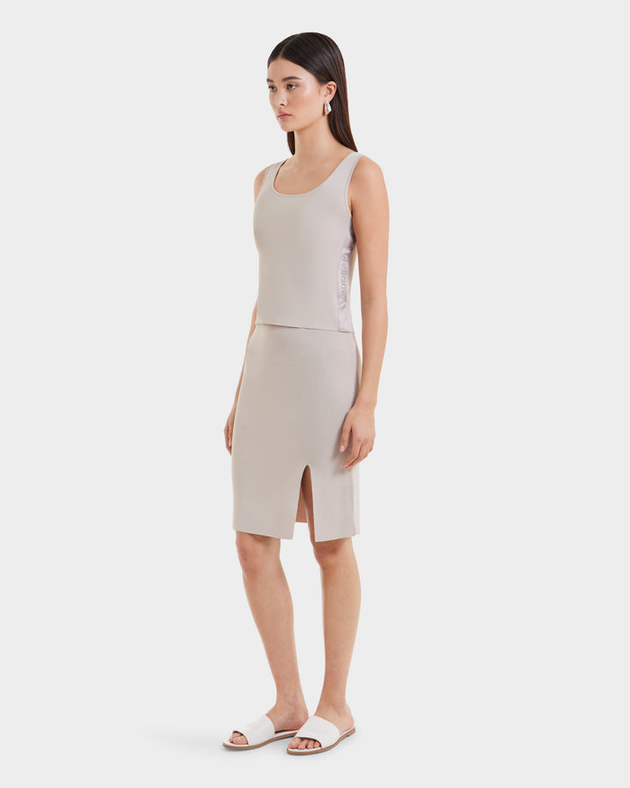 A woman wears the Rudsak MARIE - L525007 Cream sleeveless scoop-neck top and matching knee-length skirt with a front slit—an effortless, modern essential—paired with white slide sandals, straight dark hair, and drop earrings.