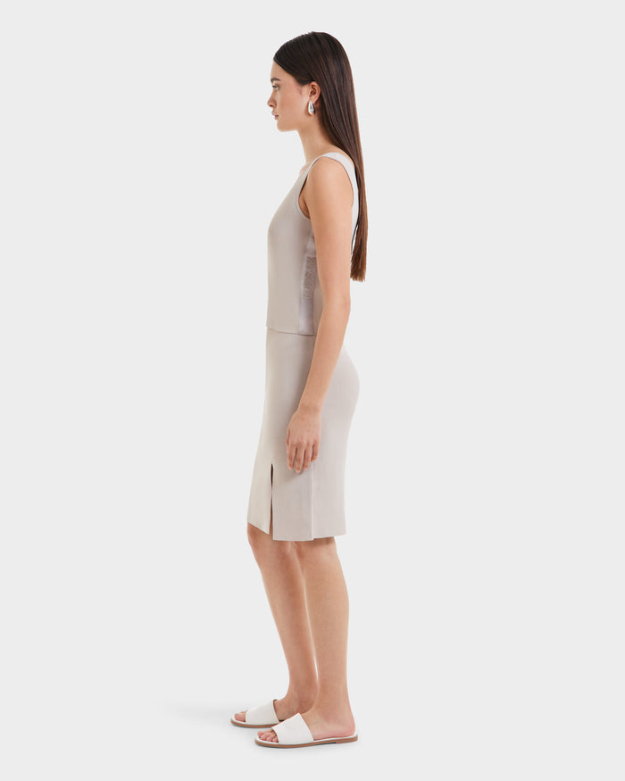 A woman with long straight hair stands in profile, wearing the Rudsak MARIE - L525007 Cream sleeveless scoop neck top and matching knee-length skirt with side slits, paired with white slide sandals against a plain white background.