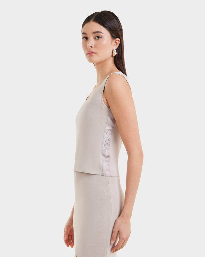 A woman with straight dark hair, dressed in Rudsak’s MARIE - L525007 Cream sleeveless knit top and matching skirt, stands in profile against a white background. The minimalist top’s side showcases subtle embossed text.