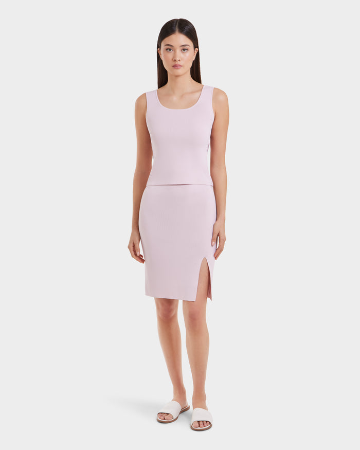 A woman stands facing forward in Rudsak’s MARIE - L525007 Light Pink sleeveless knit top and matching knee-length skirt with side slit, paired with white slide sandals against a plain white background—ideal for the modern wardrobe.