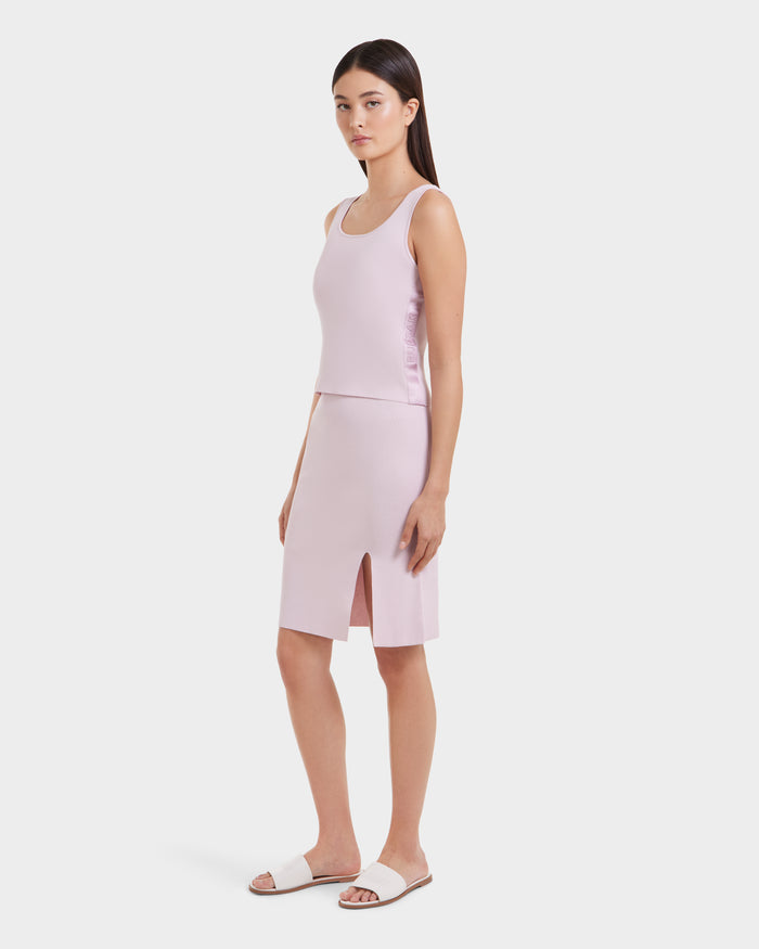 A woman poses against a plain white background wearing the Rudsak MARIE - L525007 light pink sleeveless top and matching knee-length skirt with a front slit, styled with white slide sandals for an effortlessly chic look.