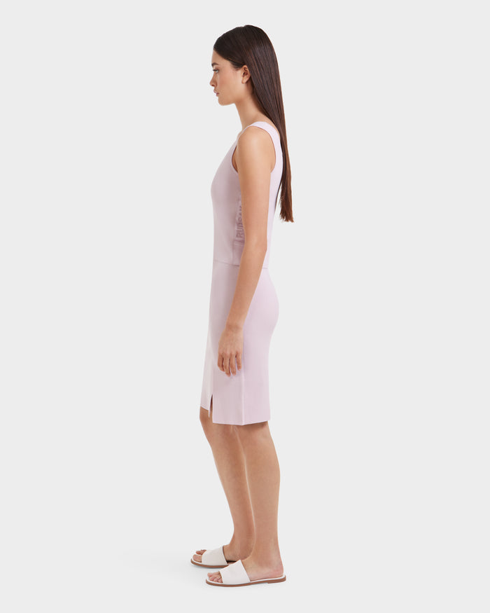A woman with long straight hair stands in profile, wearing the Rudsak MARIE - L525007 Light Pink sleeveless top and white slide sandals, creating a chic modern look against a plain white background.