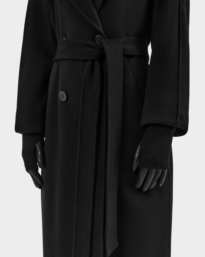 A person wears the Rudsak MARINE - 8424516 Black/Black long belted coat, styled with chic fall accessories and black women's leather gloves. The coat features a button and tie belt waist; the image is cropped from shoulders to below the knees.