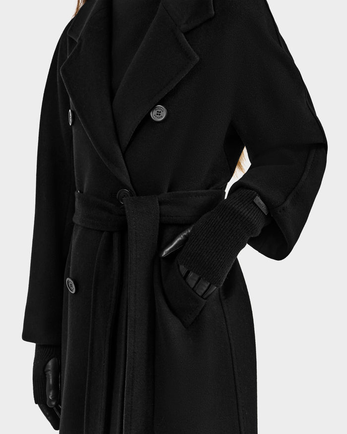 Wearing the Rudsak MARINE - 8424516 Black/Black long double-breasted coat with a fabric belt, black leather gloves, and a high-neck black top; only the lower face and upper body show against a plain light background.