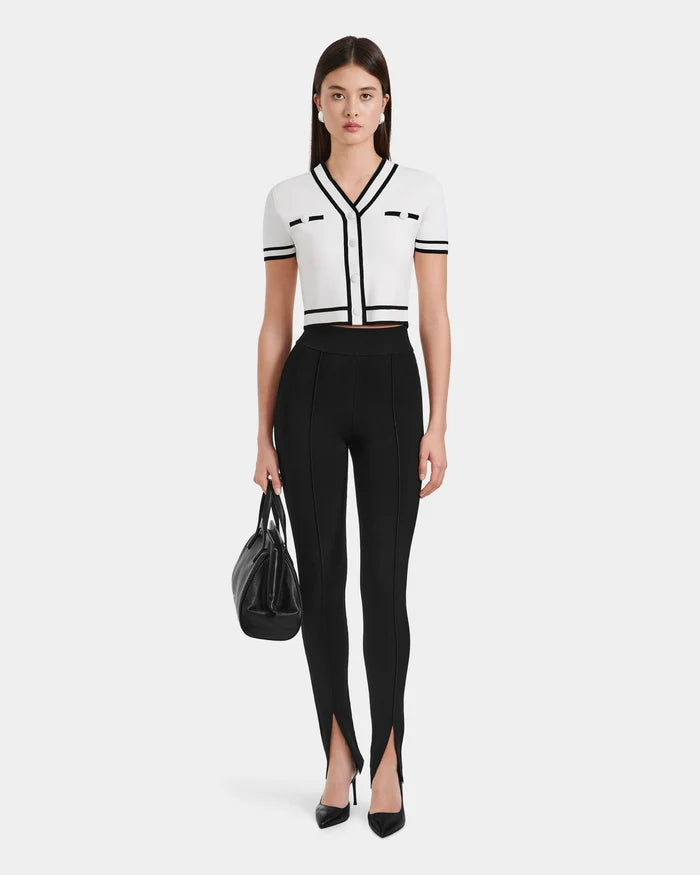A woman poses against a white background wearing a fitted white short-sleeve top with black trim, Rudsak MATHILDA - 8524515 Black front ankle slit leggings, black high heels, and holding a black handbag.