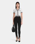 A woman poses against a white background wearing a fitted white short-sleeve top with black trim, Rudsak MATHILDA - 8524515 Black front ankle slit leggings, black high heels, and holding a black handbag.