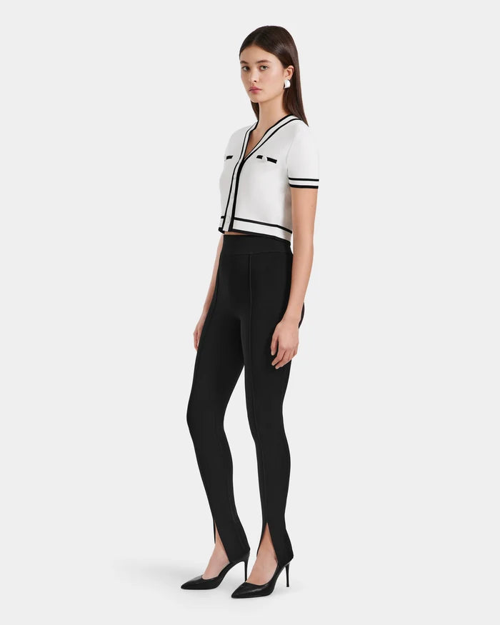A woman poses against a plain background wearing Rudsaks MATHILDA - 8524515 Black high-waisted front ankle slit leggings, paired with a white short-sleeved cropped top with black trim and black heeled shoes.