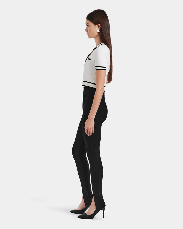 A woman in profile stands against a plain backdrop, wearing a short-sleeve white top with black trim, Rudsak’s MATHILDA - 8524515 Black high-waisted knit leggings with front ankle slits, black heels, and white earrings.