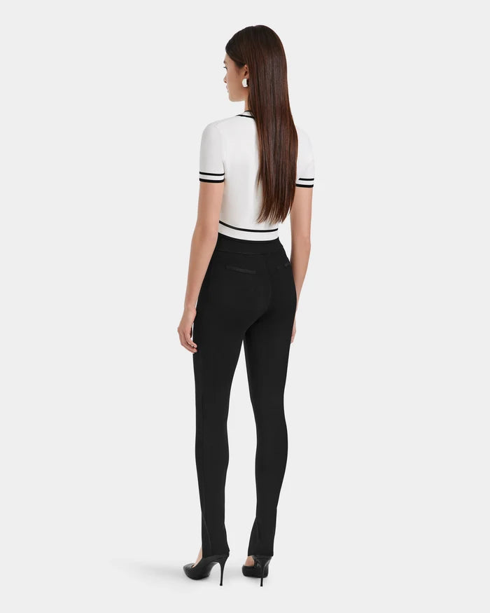 A woman with long brown hair faces away, wearing a white short-sleeve top with black trim and Rudsak MATHILDA - 8524515 Black front ankle slit leggings, paired with black heels against a plain light background.