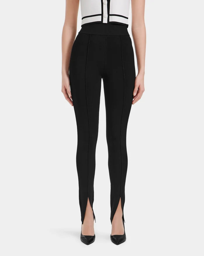 A woman faces forward in Rudsak’s MATHILDA - 8524515 Black high-waisted ankle slit leggings with vertical seams, paired with black pointed-toe heels and a white top trimmed in black.