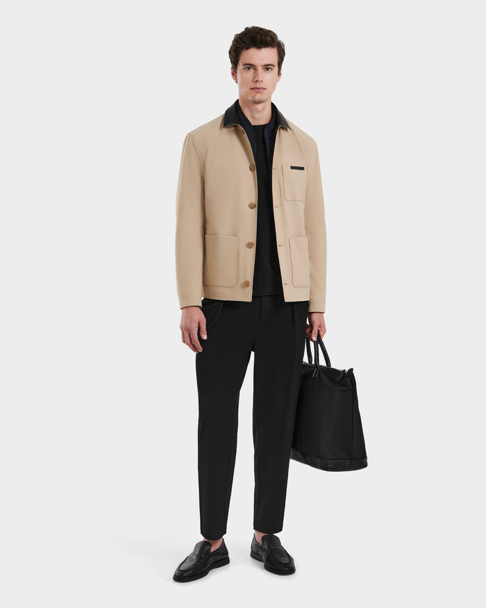 A man faces forward in Rudsaks MATTHEW - 6125004 Almond blazer-style shirt jacket, made of technical beige fabric, layered over a black shirt and pants. He holds a large black tote bag and wears black loafers against a plain white background.