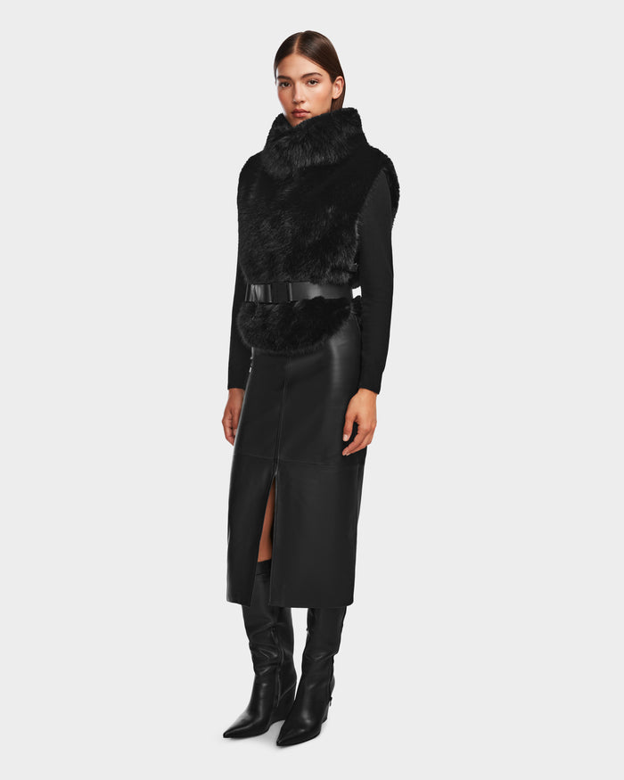 A woman wears the Rudsak MAULIN - 8524611 Black vegan leather midi skirt with a front slit, styled with a black faux fur vest, mock-neck pullover, and knee-high boots against a plain, light background.