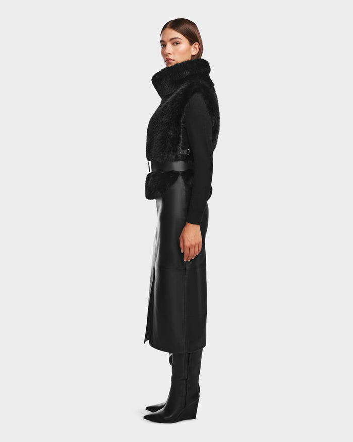 A woman stands in profile against a plain light gray background, wearing the Rudsak MAULIN - 8524611 Black belted vegan leather midi skirt styled with a black faux fur vest, black mock-neck pullover, and knee-high boots.