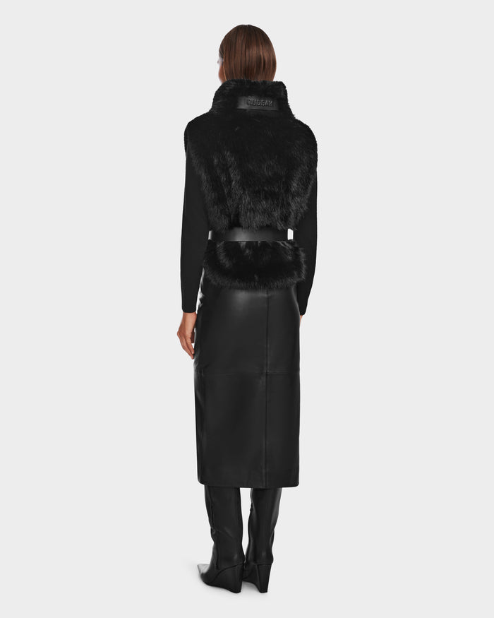 A person with straight brown hair stands facing away, wearing the Rudsak MAULIN - 8524611 Black faux fur vest, a black mock-neck pullover, a vegan leather midi skirt, and tall black boots against a plain light background.