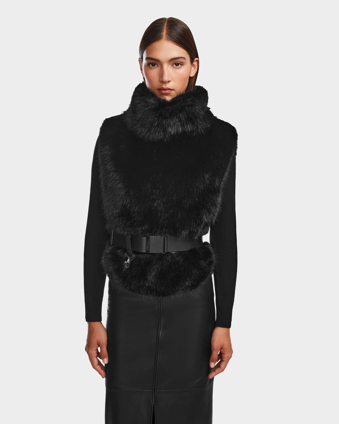 A woman with straight brown hair models the Rudsak MAULIN - 8524611 Black high-neck faux fur vest, belted at the waist, over a black top and skirt, standing against a plain light background.