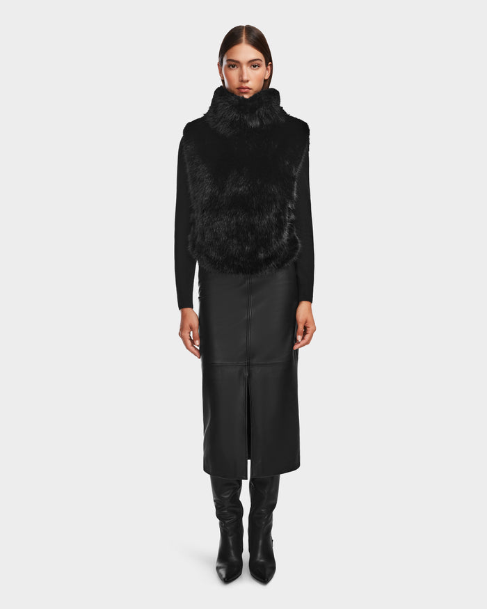 A woman with straight brown hair and a neutral expression models the Rudsak MAULIN - 8524611 Black faux fur vest over a long vegan leather skirt with a front slit and knee-high black boots against a plain background.