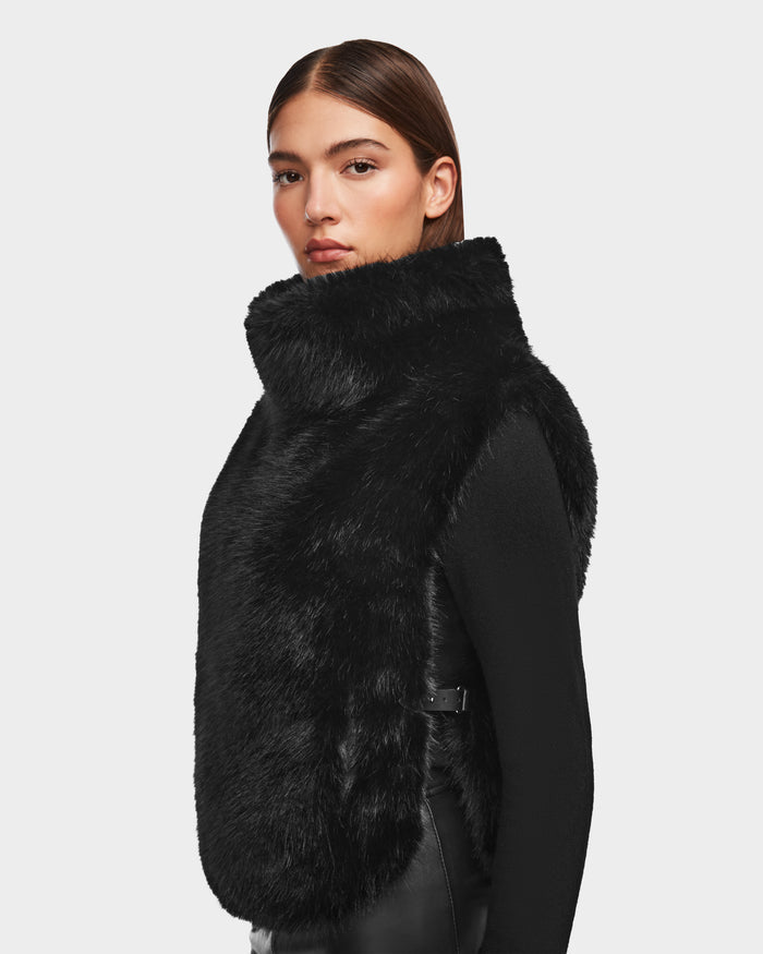 A woman with straight brown hair models the Rudsak MAULIN - 8524611 Black faux fur vest over a black long-sleeve top, standing against a plain light background and gazing slightly past the camera.