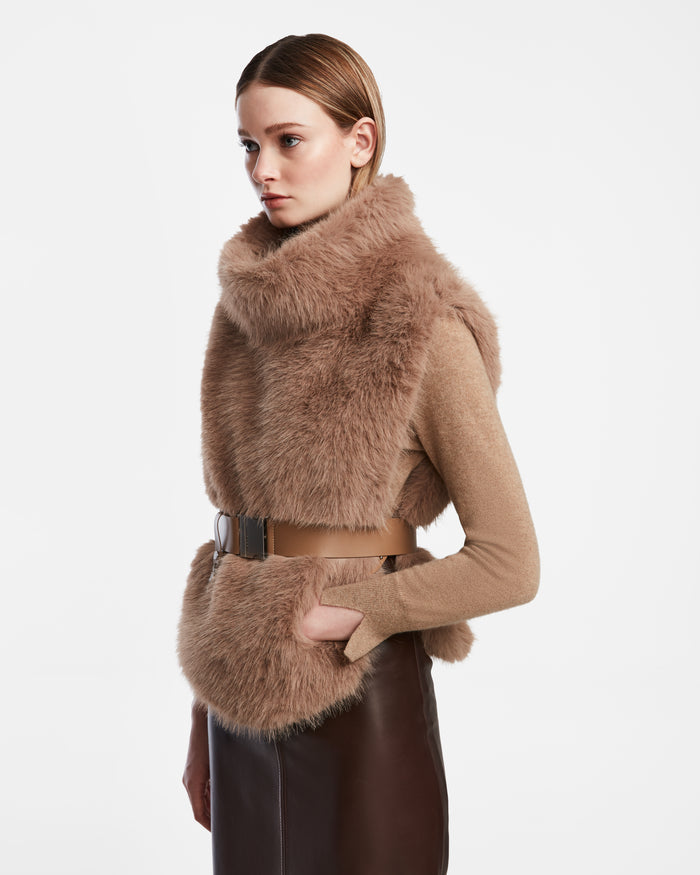 A woman wears the Rudsak MAULIN - 8524611 Camel faux fur vest with a wide tan belt over a beige long-sleeve top and dark brown skirt, standing against a plain white background.