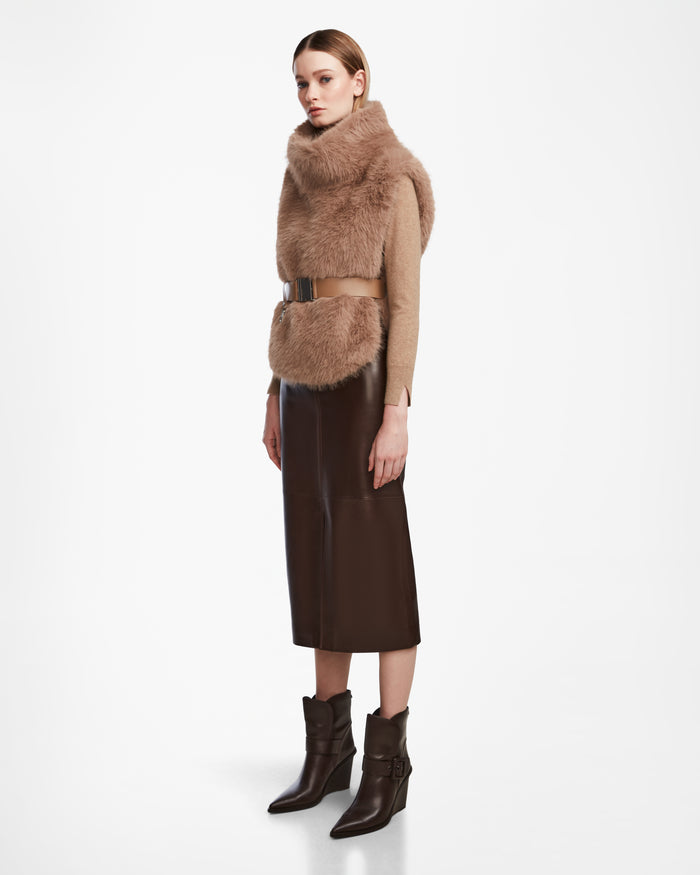 A woman poses against a white background in the Rudsak MAULIN - 8524611 Camel faux fur vest, styled over a tan mock-neck pullover with a dark brown vegan leather midi skirt, metallic belt, and brown ankle boots.