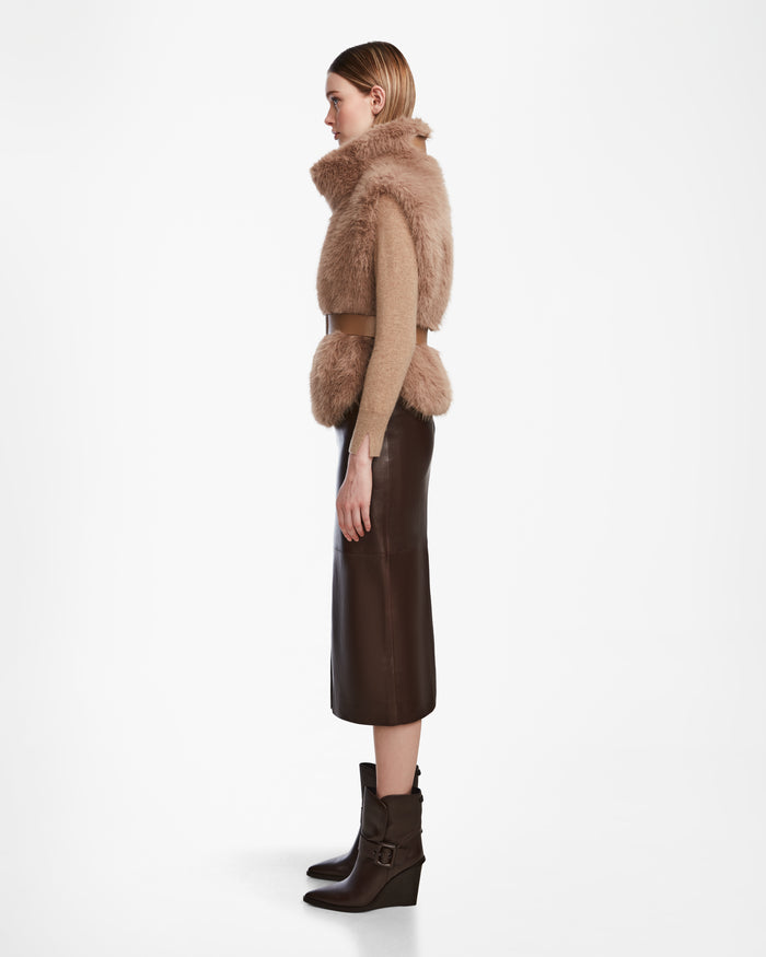 A woman stands in profile wearing the Rudsak MAULIN - 8524611 Camel faux fur vest over a beige mock-neck pullover, paired with a dark brown midi skirt and matching buckle ankle boots, set against a plain white background.