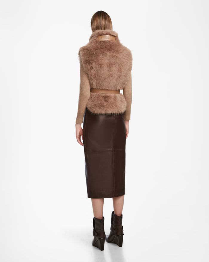 A person faces away, wearing the Rudsak MAULIN - 8524611 Camel faux fur vest over a tan mock-neck pullover, paired with a dark vegan leather midi skirt and brown pointed-toe ankle boots. Straight hair falls just past the shoulders.