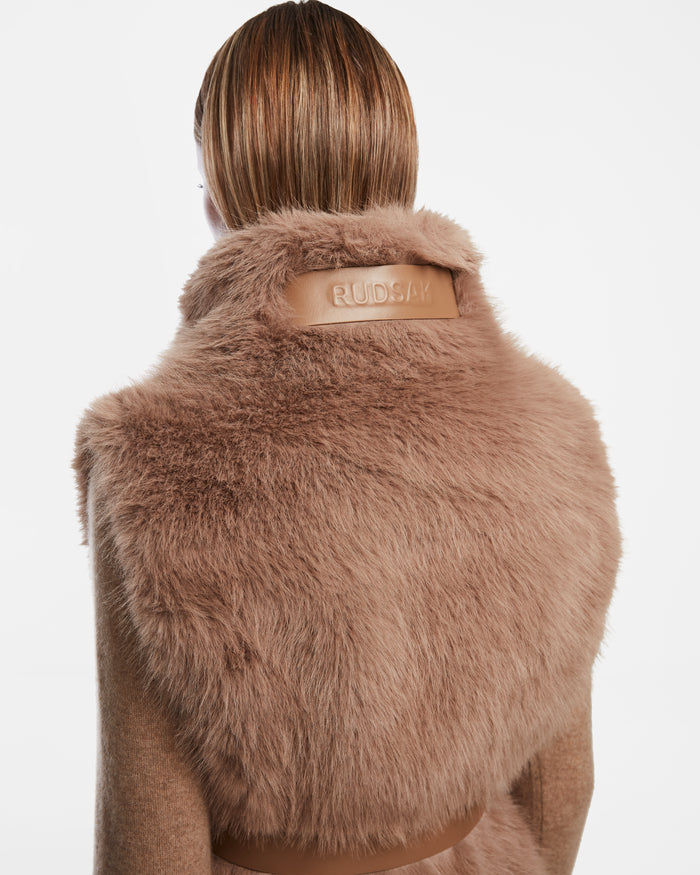 Seen from behind, a person with straight, light brown hair wears the Rudsak MAULIN - 8524611 Camel—a soft brown faux fur vest featuring a vegan leather collar patch embossed with Rudsak branding. The background is plain white.