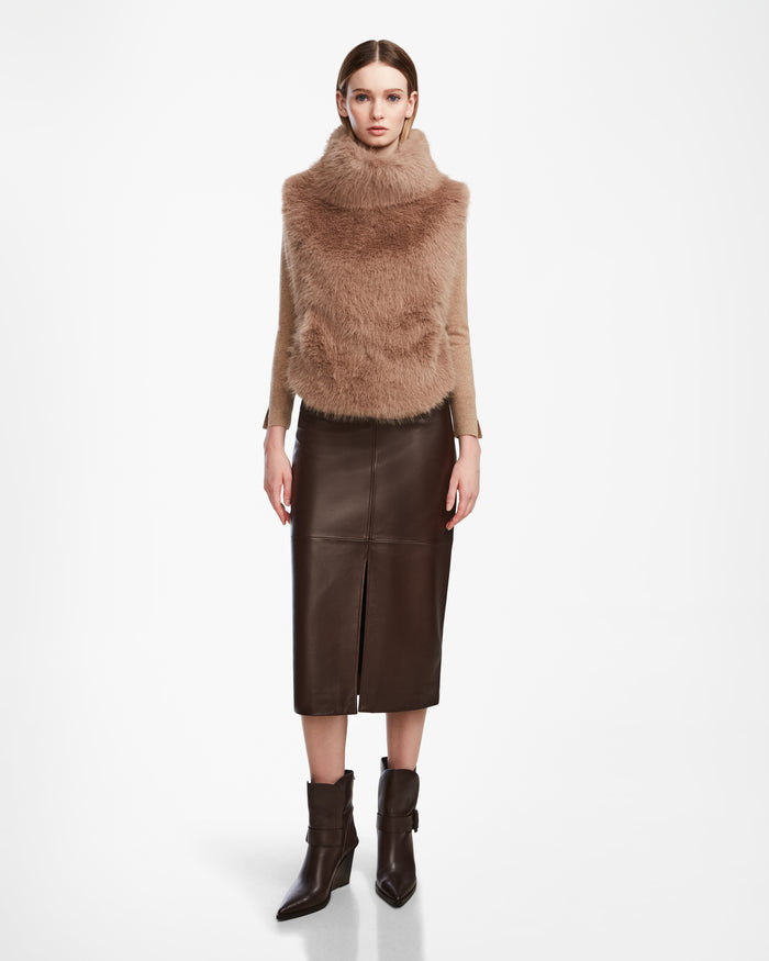 A woman stands against a white background wearing the MAULIN - 8524611 Camel faux fur vest by Rudsak, paired with a long brown vegan leather skirt with a front slit and dark brown ankle boots.