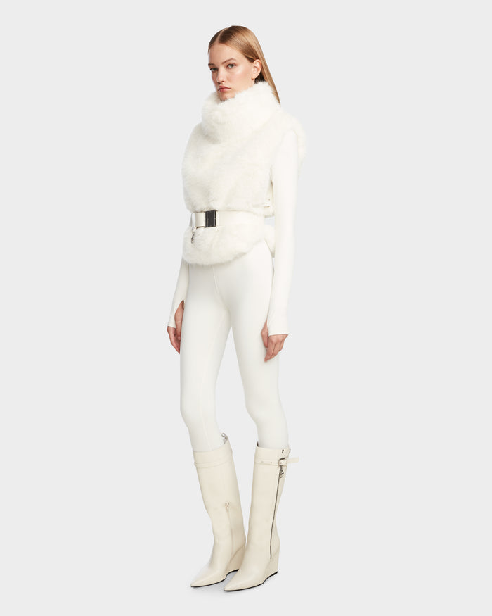 A woman in Rudsaks MAULIN - 8524611 Iceland all-white outfit—faux fur vest, metallic belt, vegan leather pants, and knee-high heeled boots—poses against a light gray background.
