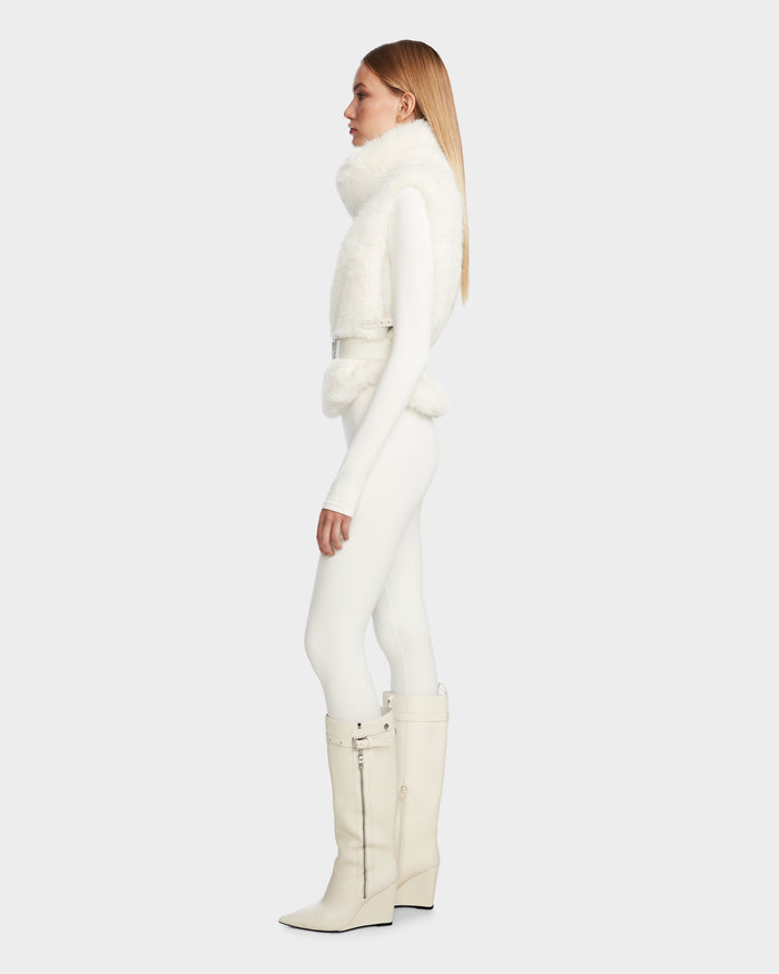 A woman with long, straight light brown hair in profile wears the Rudsak MAULIN - 8524611 Iceland: an all-white outfit with a fluffy faux fur vest, fitted pants, and knee-high boots against a plain light background.
