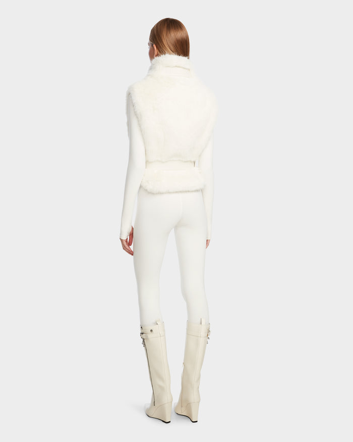 A model with long, straight hair wears Rudsak’s MAULIN - 8524611 Iceland sleeveless faux fur vest over a fitted white outfit and knee-high cream boots, shown from the back against a plain gray background.
