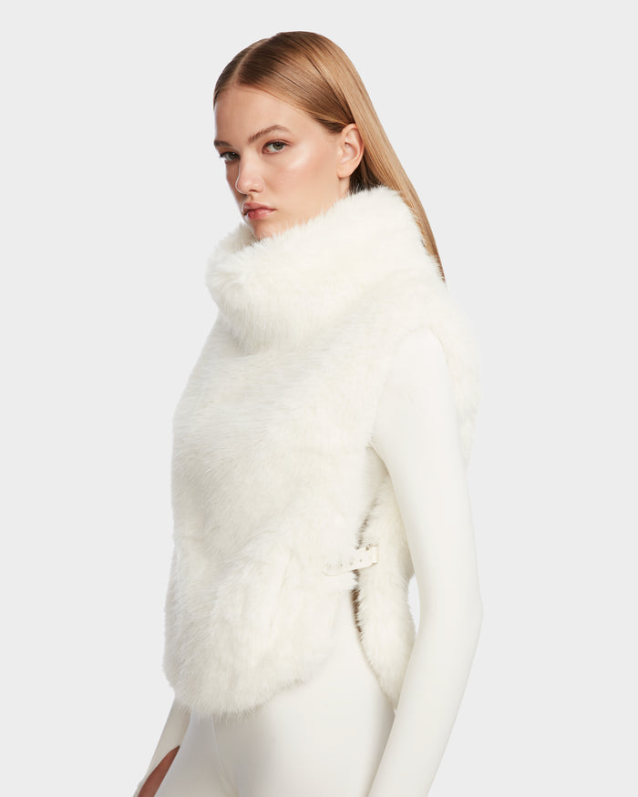 A woman with straight, light brown hair models the Rudsak MAULIN - 8524611 Iceland white faux fur vest with side buckles over a fitted white mock-neck pullover, standing against a plain light gray background.