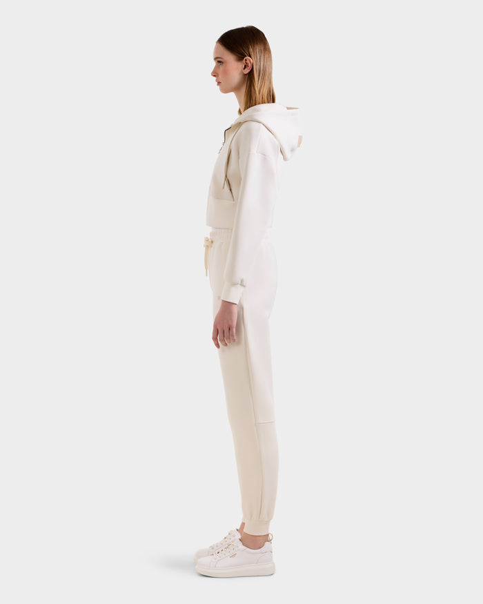 A woman stands in profile against a white background, wearing the Rudsak MAXTON - U525007 Vanilla W jogger set with matching hoodie and white sneakers for everyday comfort.