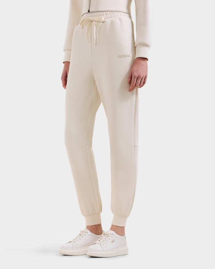 A person wears the Rudsak MAXTON - U525007 Vanilla W jogger pants—cream-colored, cotton-blend unisex joggers with an elastic waistband, drawstring, and subtle branding—styled with a matching zip-up top and white sneakers against a light background.