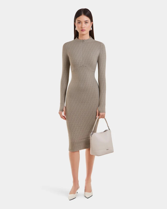 A woman faces forward in the Rudsak MAYLIN - 8523504 Cement Pattern dress, styled with white pointed shoes and a light beige handbag. She has straight brown hair, a neutral expression, and stands against a plain white background.
