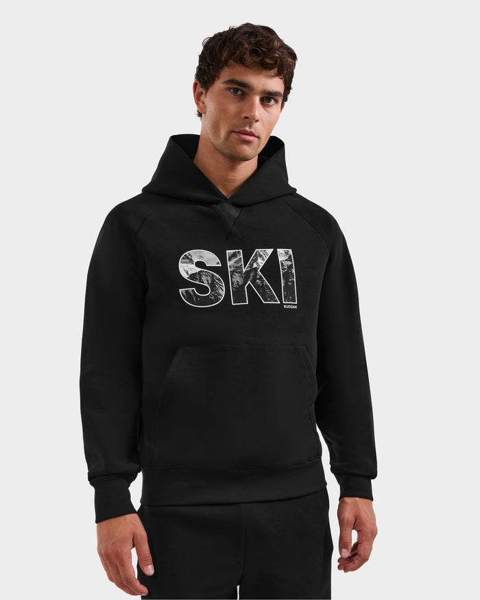 A man wears the Rudsak MIKAEL - 6525647 Black men’s ski hoodie featuring bold, textured white "SKI" lettering on the front, standing against a plain light gray background.