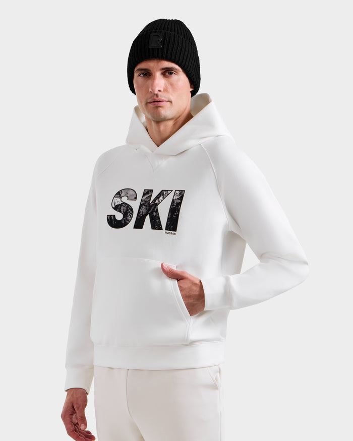 A man wears the Rudsak MIKAEL - 6525647 Iceland white men's ski hoodie featuring "SKI" on the front and a black knit beanie, standing against a plain light background with one hand in his pocket.