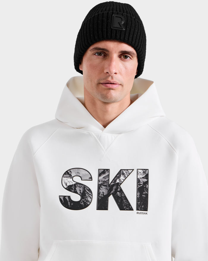 A man wearing a black knit beanie and the Rudsak MIKAEL - 6525647 Iceland premium cotton men’s ski hoodie with bold “SKI” text stands against a plain light background, looking directly at the camera.