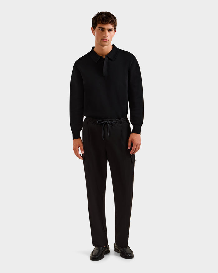 A man faces forward with a neutral expression, wearing Rudsak’s MIKEY - 6525597 Black long-sleeve polo, tailored premium trousers, and black shoes against a plain light background.