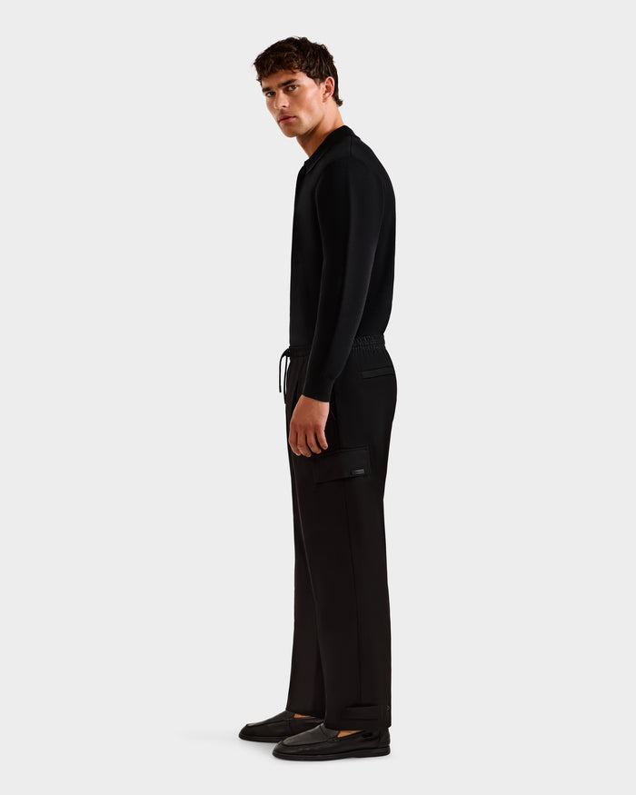 A man stands sideways against a plain light background, wearing Rudsak's MIKEY - 6525597 Black long-sleeve shirt, tailored men's trousers with a side pocket, and black shoes.
