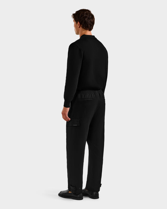 A person faces away from the camera, wearing Rudsak MIKEY - 6525597 Black men's tailored fit trousers with a black long-sleeve top and black shoes against a light gray background.