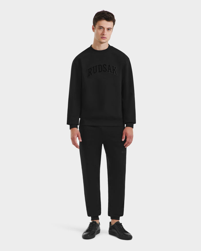 A young man faces forward against a white background, wearing the Rudsak MILOS - 6525646 Black men’s premium cotton blend crewneck sweatshirt with matching black pants and sneakers.