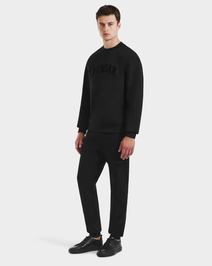 A man stands against a white background, wearing the Rudsak MILOS – 6525646 Black men’s premium cotton blend crewneck sweatshirt with matching black jogger pants and black sneakers.