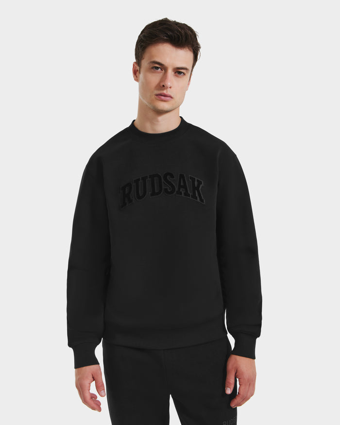 A young man stands against a white background, wearing Rudsak’s MILOS - 6525646 Black sweatshirt and black pants. The premium crewneck features “RUDSAK” in bold, embossed letters across the chest.